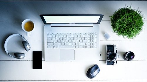 Bild mit aufgeklapptem Laptop, Kopfhörer, Tasse Kaffee, Handy, Fotoapparat, und grüne Pflanze